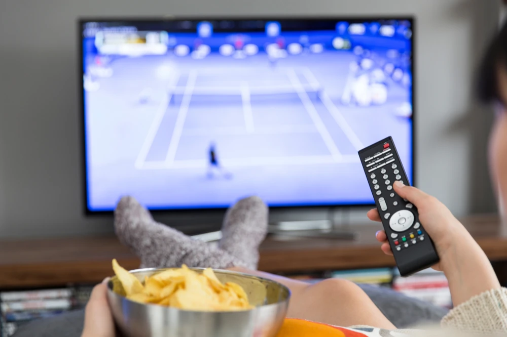 persoon met remote in de hand en schaal chips op schoot, televisie kijkt.