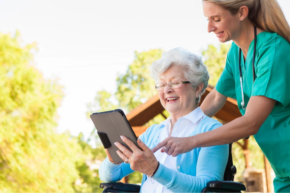 oudere dame in rolstoel met iPad in haar hand, wordt geholpen door verpleegkundige.